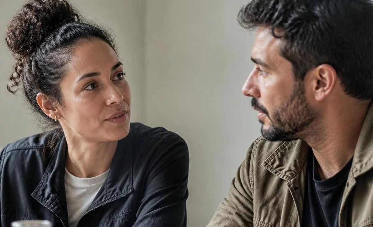 Três participantes conversam ao redor de uma mesa comprida numa sala clara de janelas altas